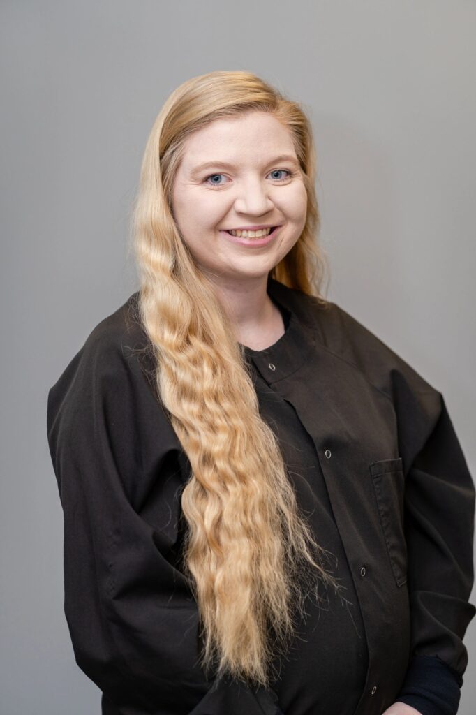 A woman with long blonde hair standing in front of a gray background.